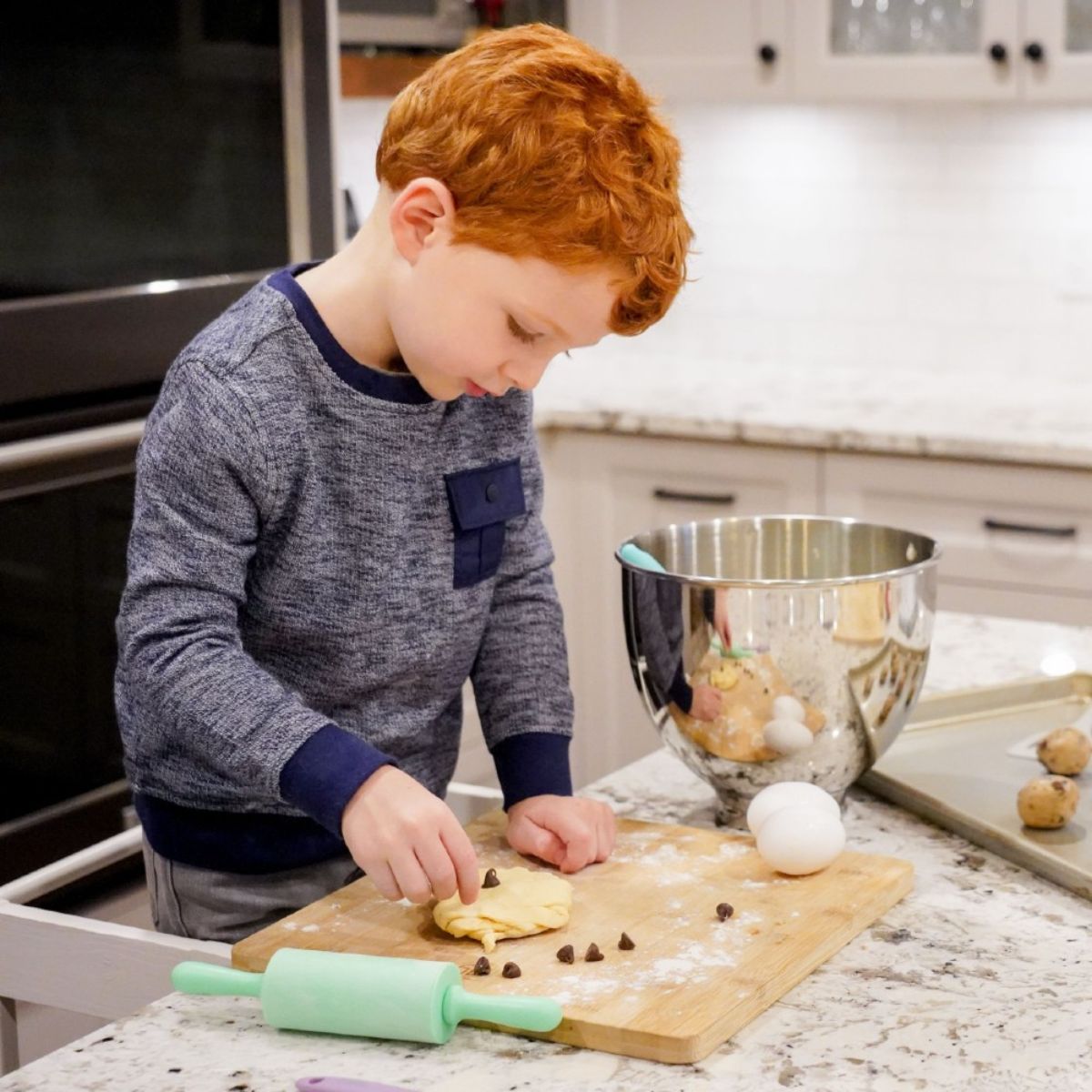 Child in a kitchen preparing food on a cutting board with a rolling pin and ingredients.
