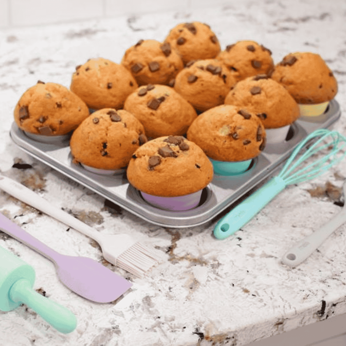 A tray of chocolate chip muffins on a white marble-like countertop with baking utensils nearby, including a whisk, spatula, brush, and rolling pin.