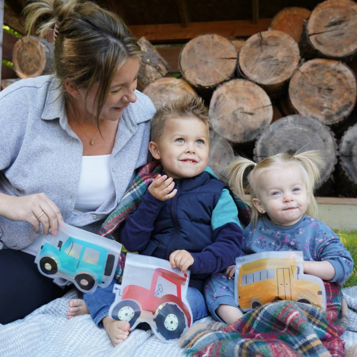 Woman and two children sitting on a blanket with toy vehicles, surrounded by wooden logs.