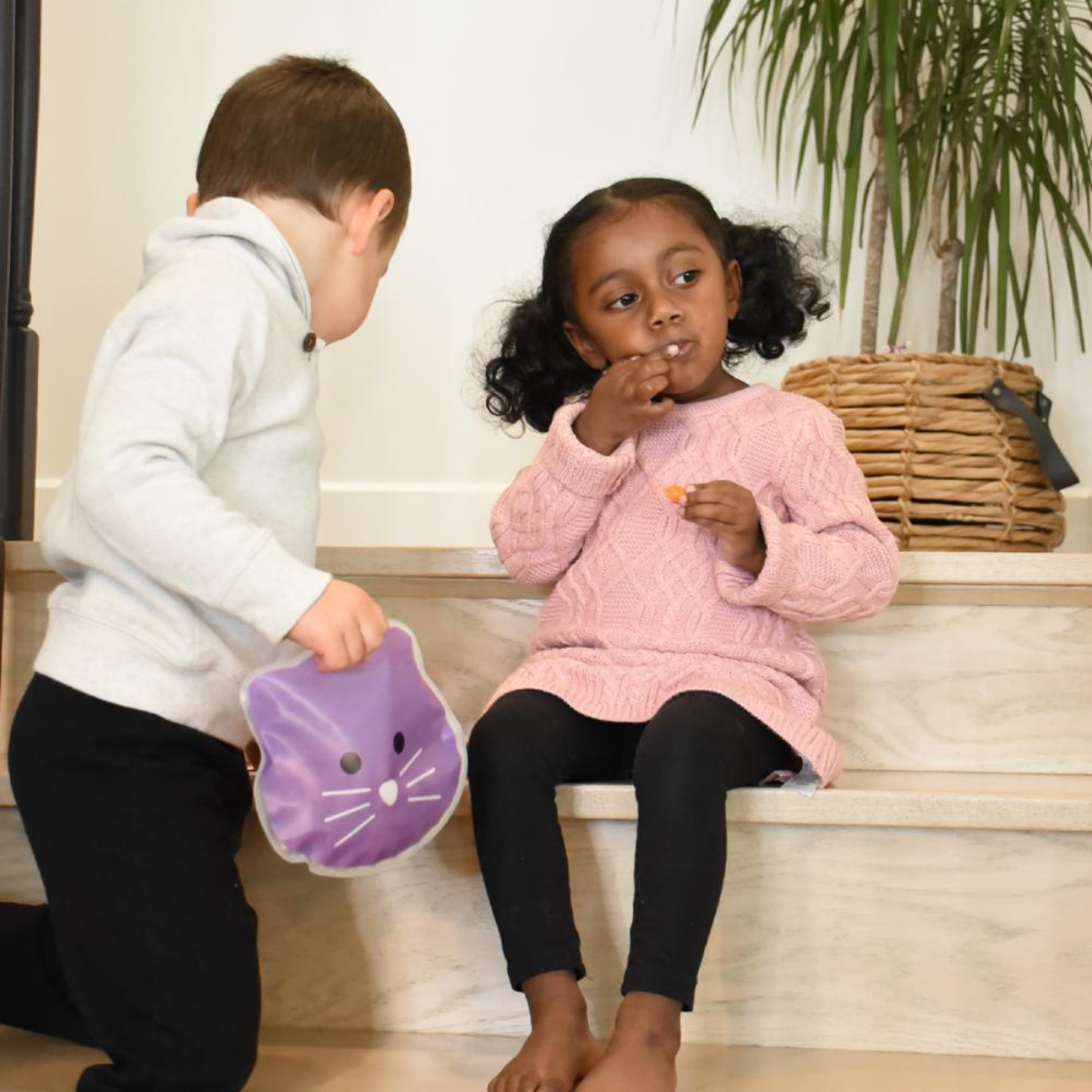 Two children sitting on a step indoors with a purple cat-shaped snackbag.