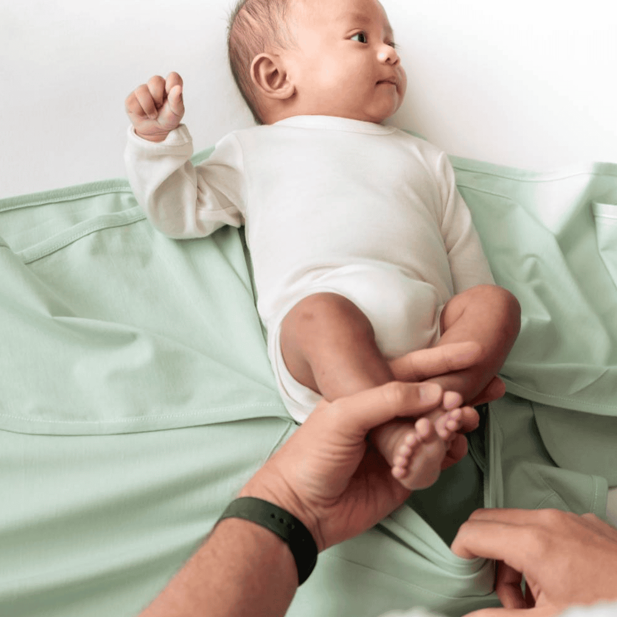 Baby lying on a light green blanket with a person's hand holding the baby's foot.