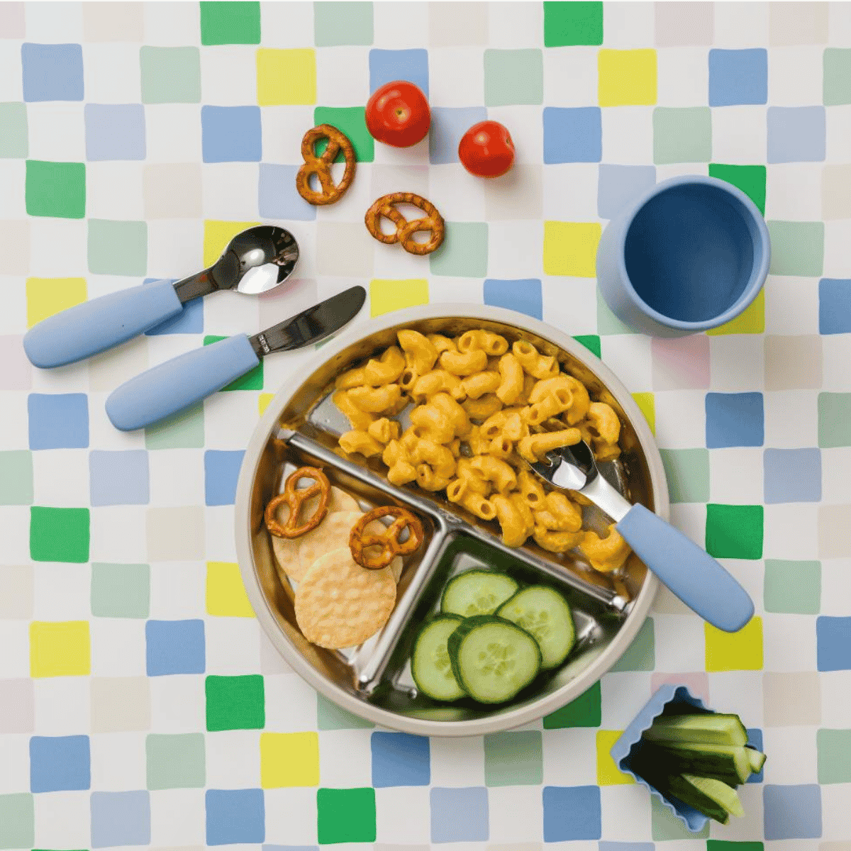 A colorful children's divided lunch tray filled with macaroni, cucumber slices, crackers, and pretzels, styled on a pastel checkered tablecloth with blue-handled utensils and a blue cup.
