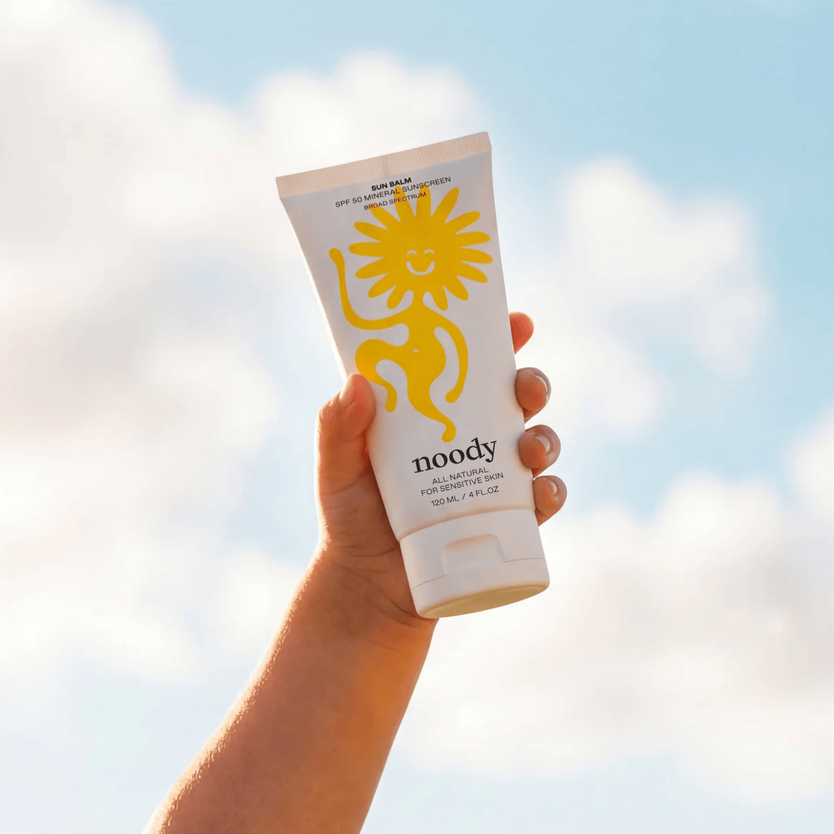 Hand holding a noody sunscreen tube against a bright sky