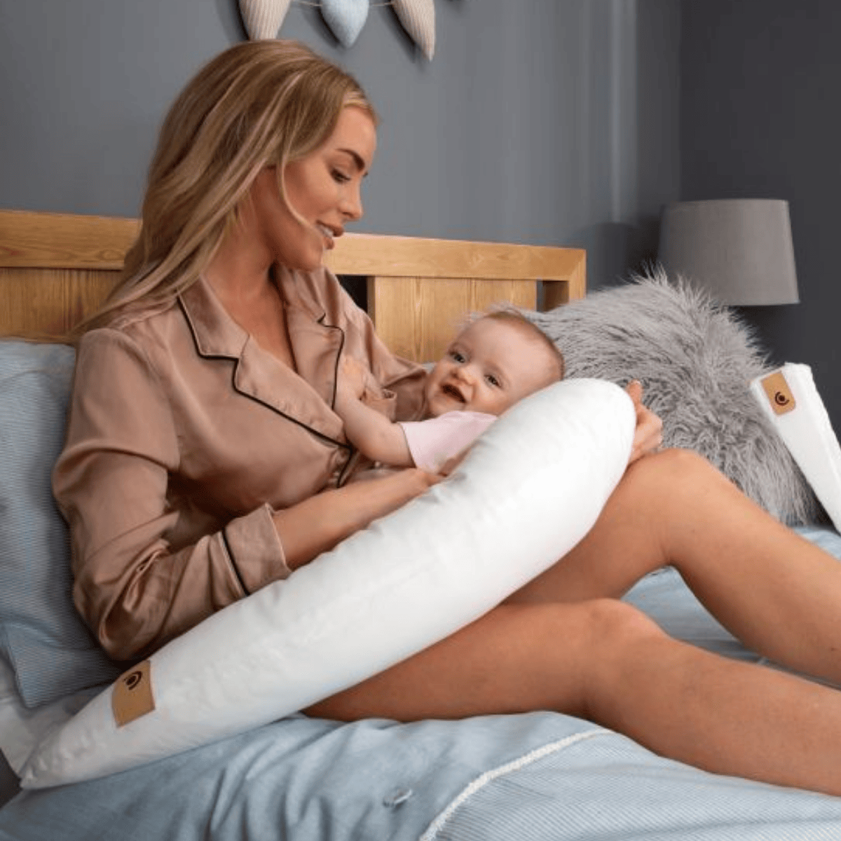 Woman in pajamas holding a baby with a U-shaped pillow on a bed.