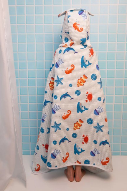 Person wrapped in a colorful hooded towel with sea creature pattern against a blue tiled wall.
