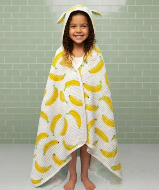 Child wrapped in a banana-patterned hooded towel against a tiled wall background