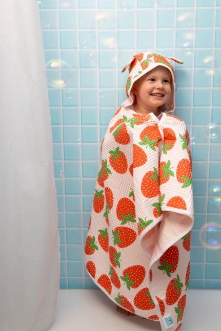 Child wrapped in a strawberry-patterned hooded towel against a tiled bathroom wall.
