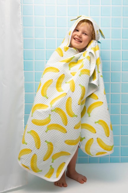 Child wrapped in a banana-patterned hooded towel against a blue tiled wall.