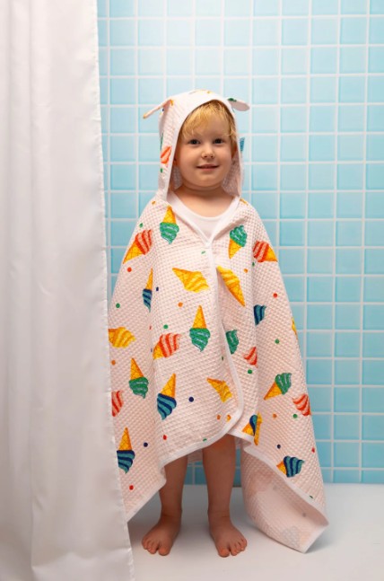 Child wearing a colorful hooded towel with icecram patterns in a bathroom.