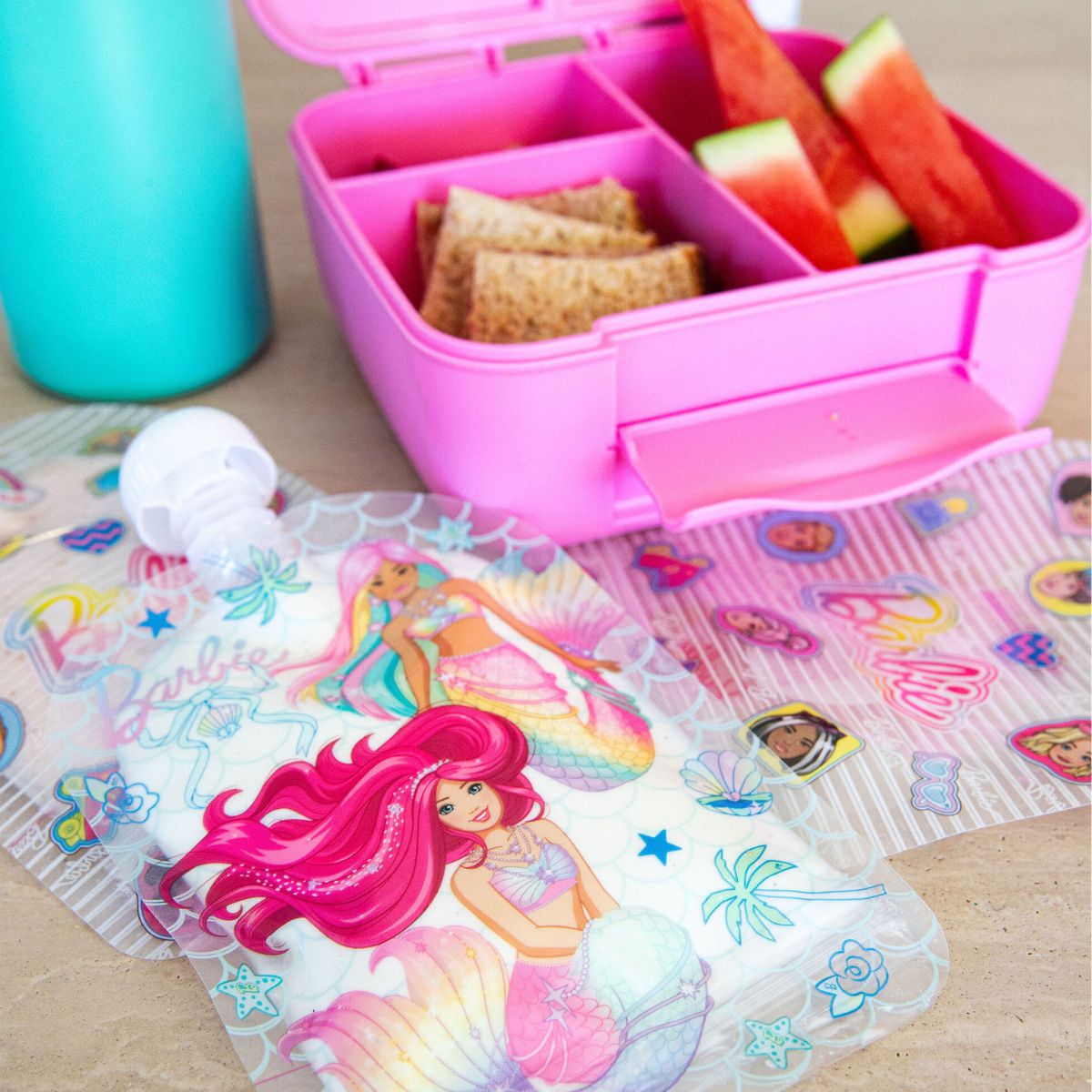 Pink bento box with snacks and a water bottle on a table with a Barbie-themed sinchie.