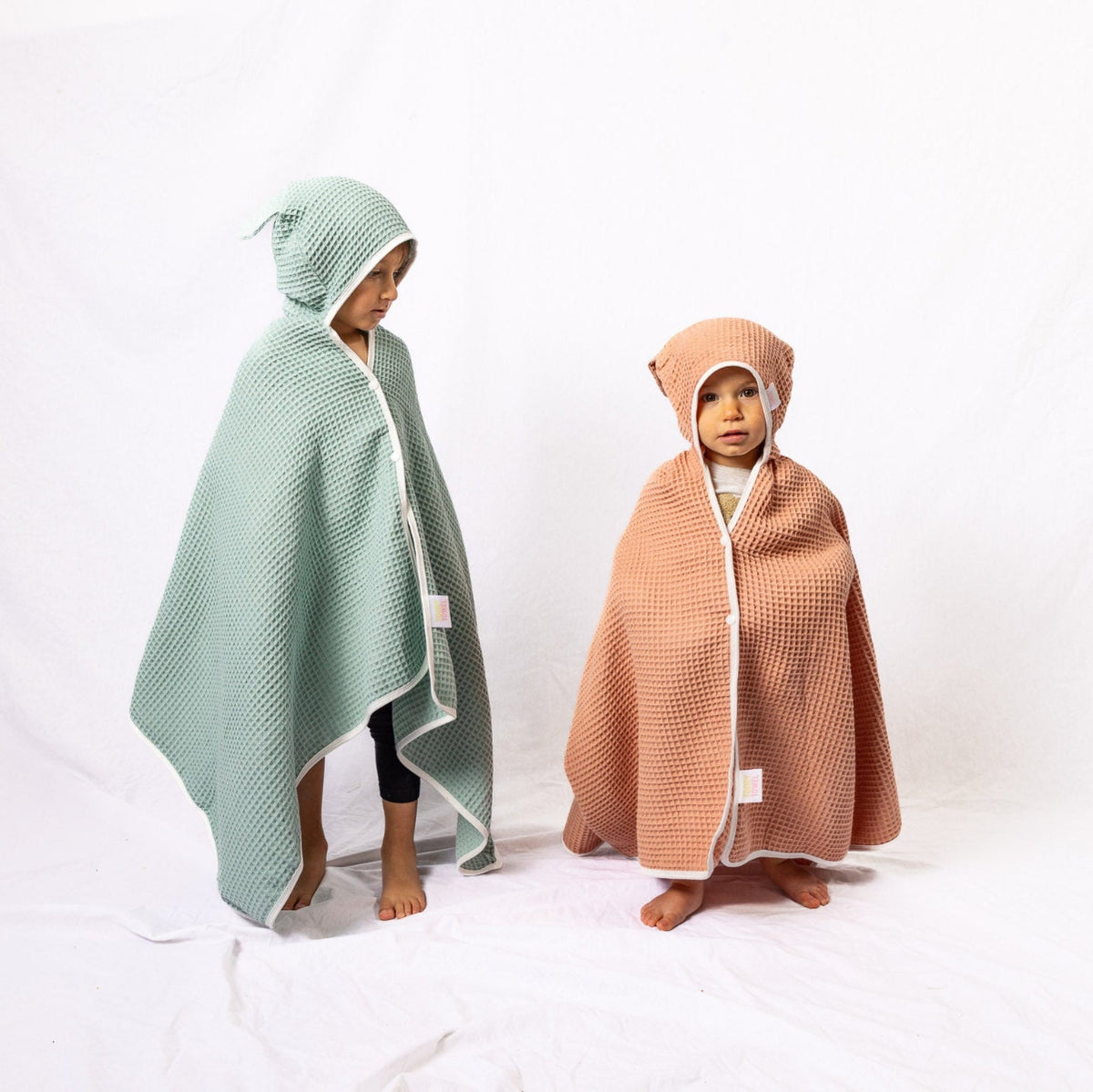 Two children wearing hooded blankets, one green and one brown, on a white background.