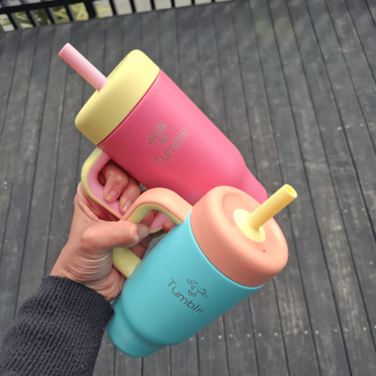 Two colorful tumblers with straws held by a person on a wooden deck.