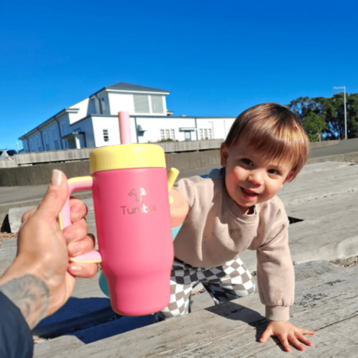 Child holding a pink tumbler with a yellow lid outdoors on a sunny day.