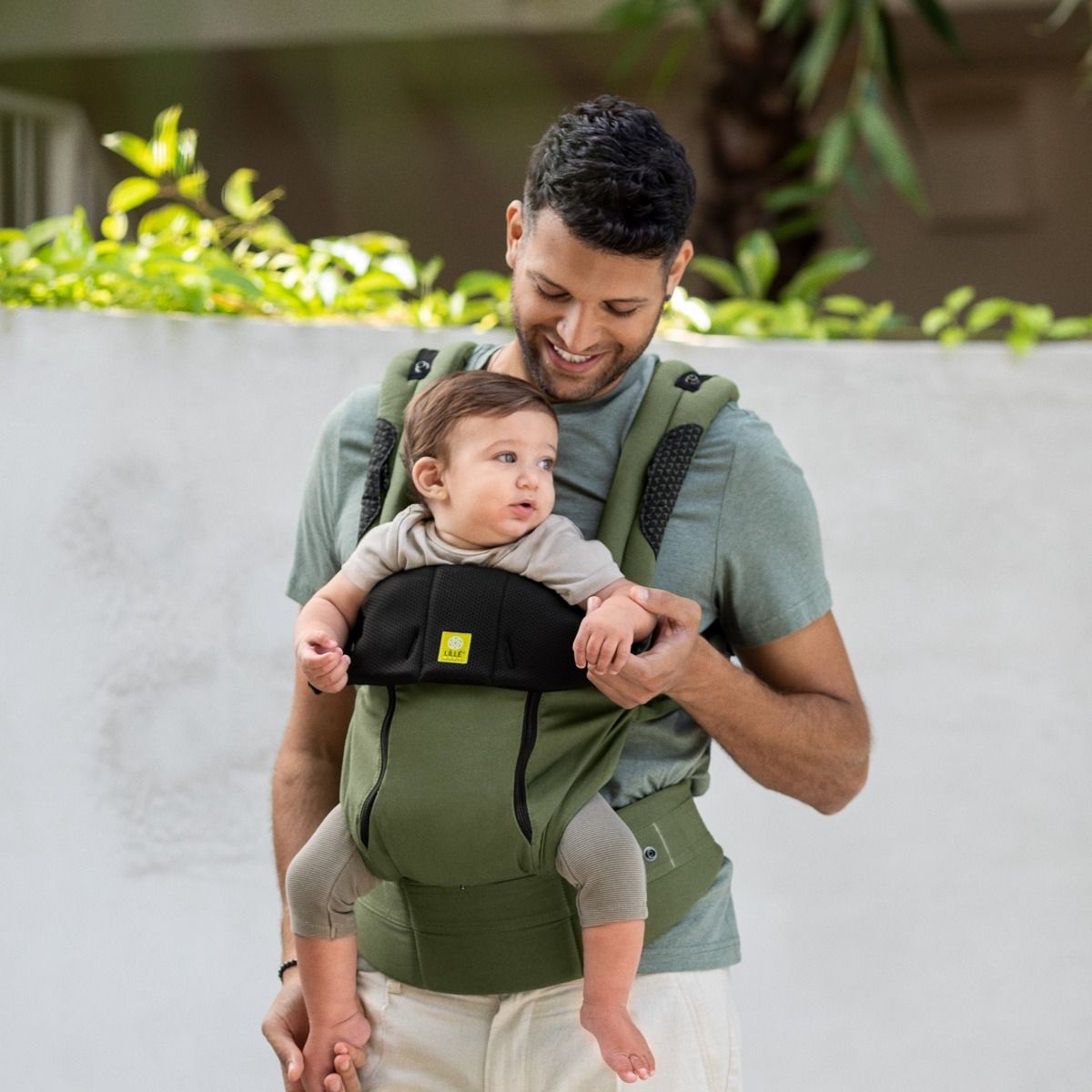 Man carrying a baby in a olive green baby carrier against a white wall with greenery.