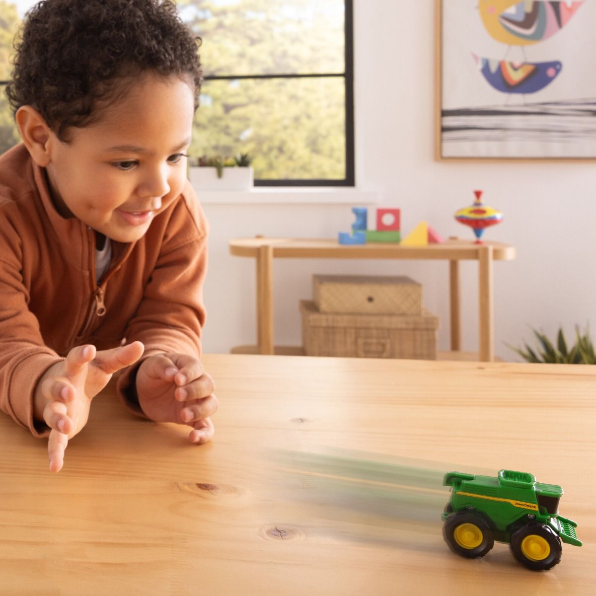 Child playing with a John Deere toy tractor on a wooden table in a room with toys and a window in the background.