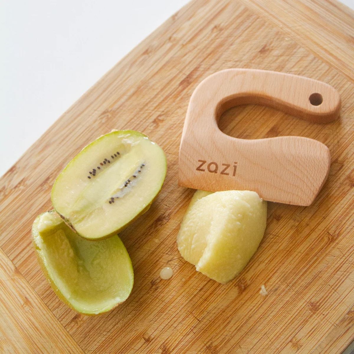 Wooden knife with &#39;zazi&#39; branding on a wooden cutting board with a halved kiwi and a piece of peeled apple.